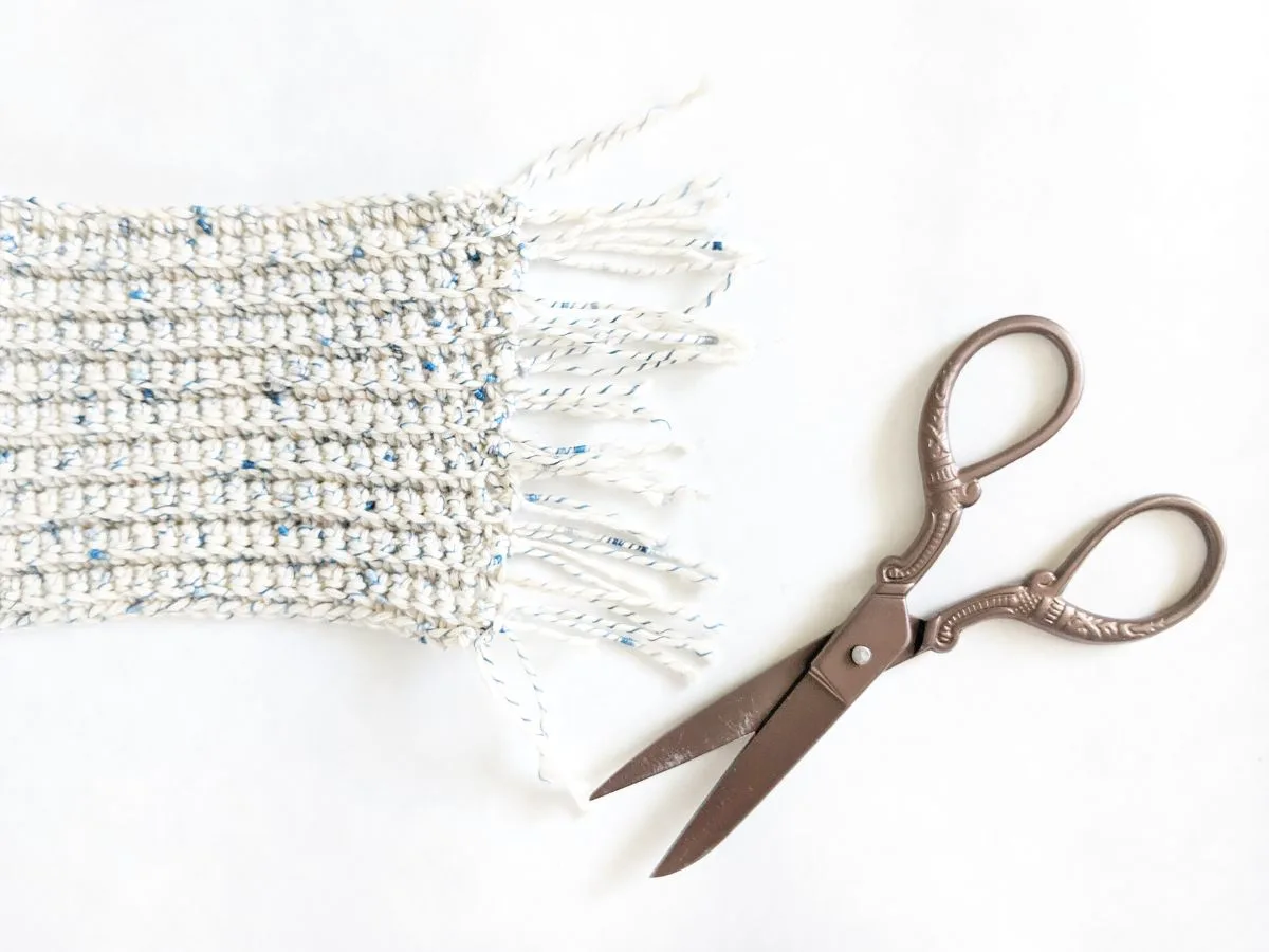 A cotton speckled crochet mug rug coaster with fringe and a pair of scissors. 