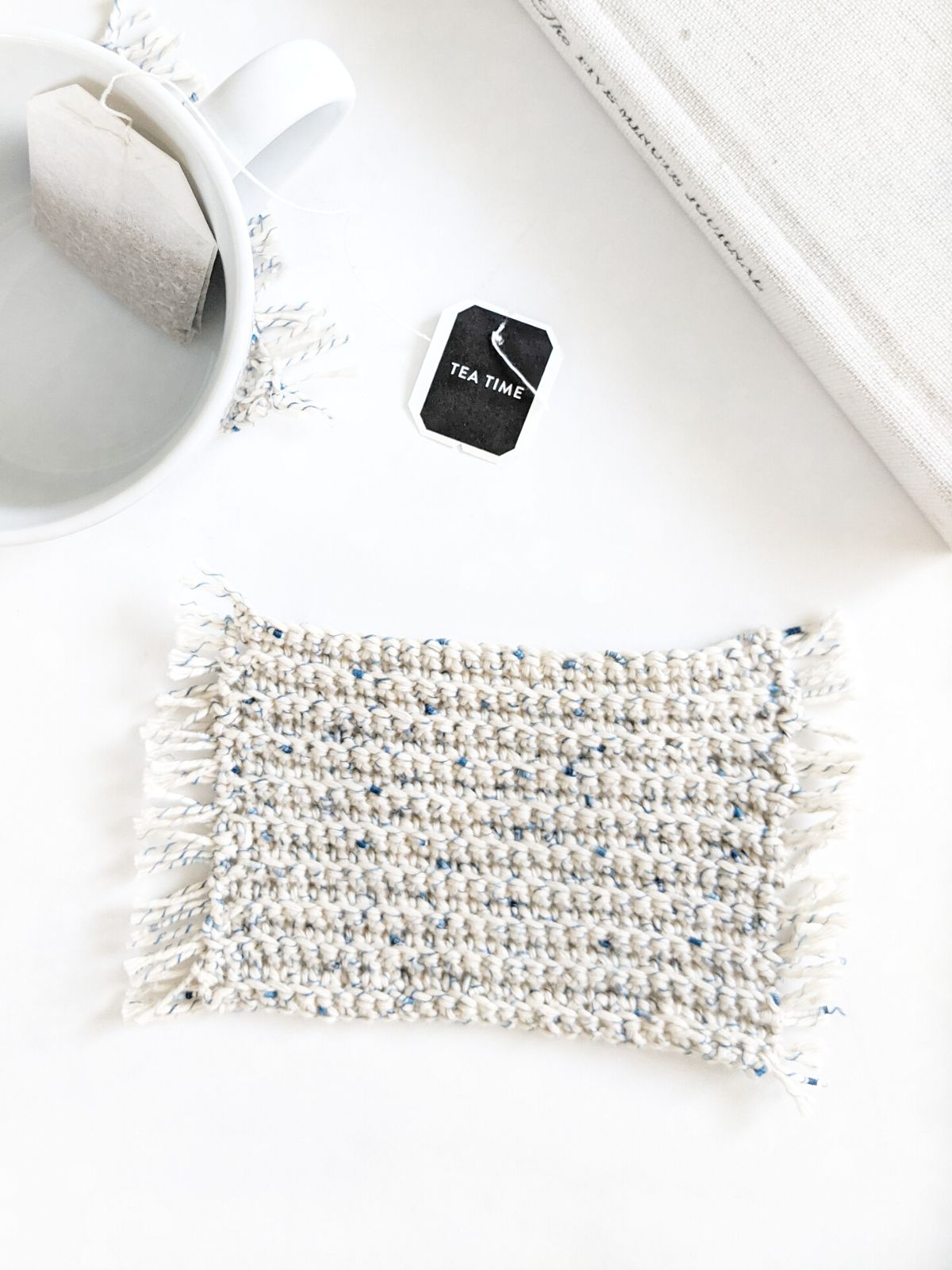A crochet coaster with fringe and a teacup with a tea bag. 