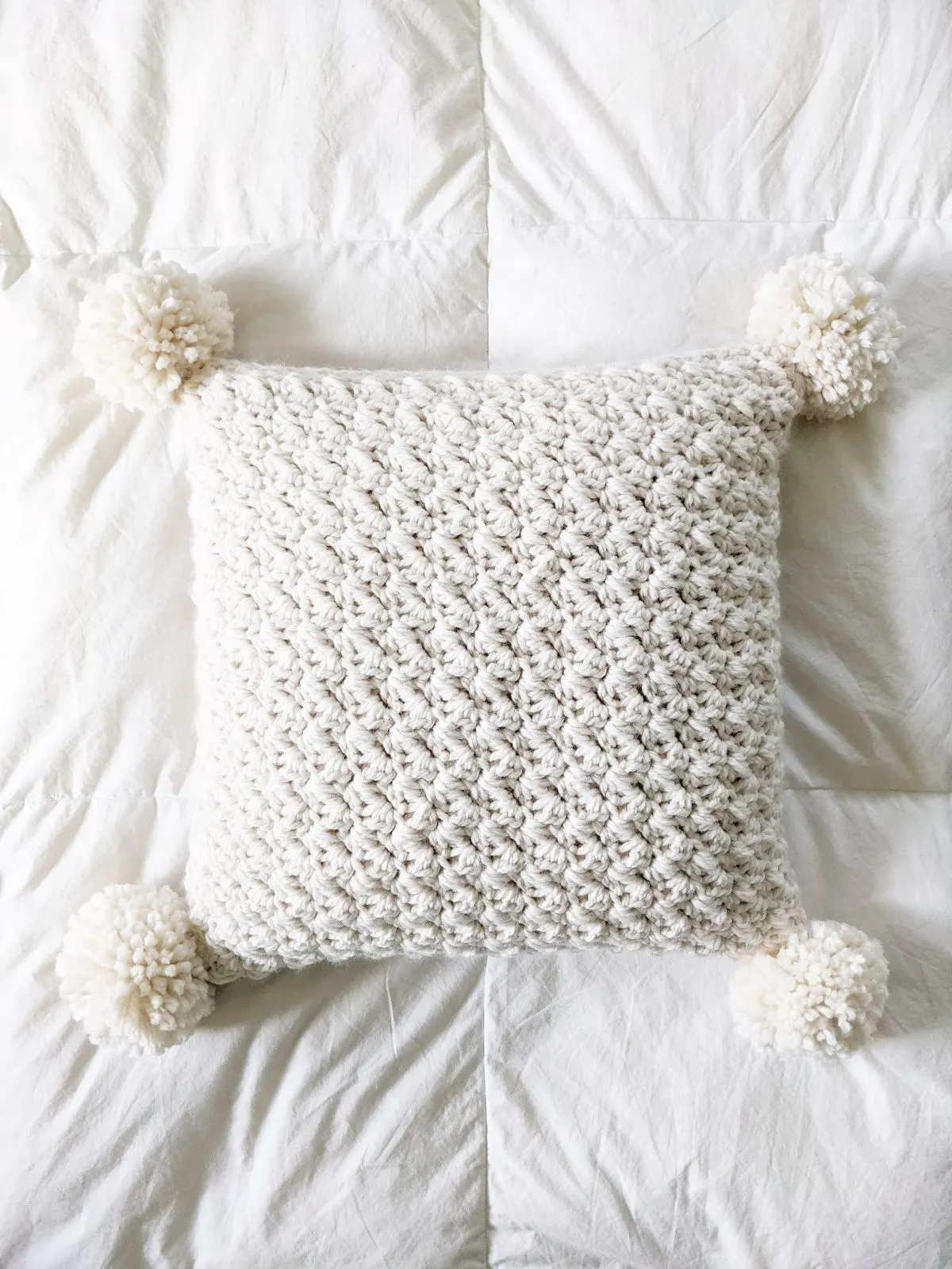 a chunky crochet pillow with pom-poms on a white ikea comforter