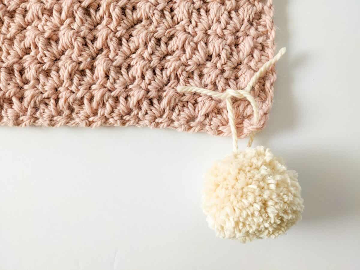 Image shows step 2 in attaching a pom pom to your crochet blanket.