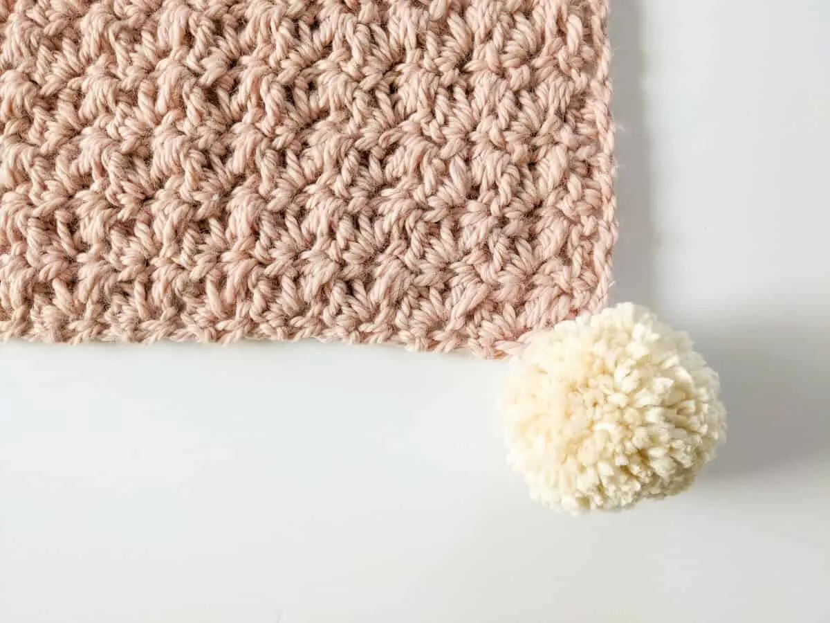 Image shows a crochet blanket with a large pom pom. 