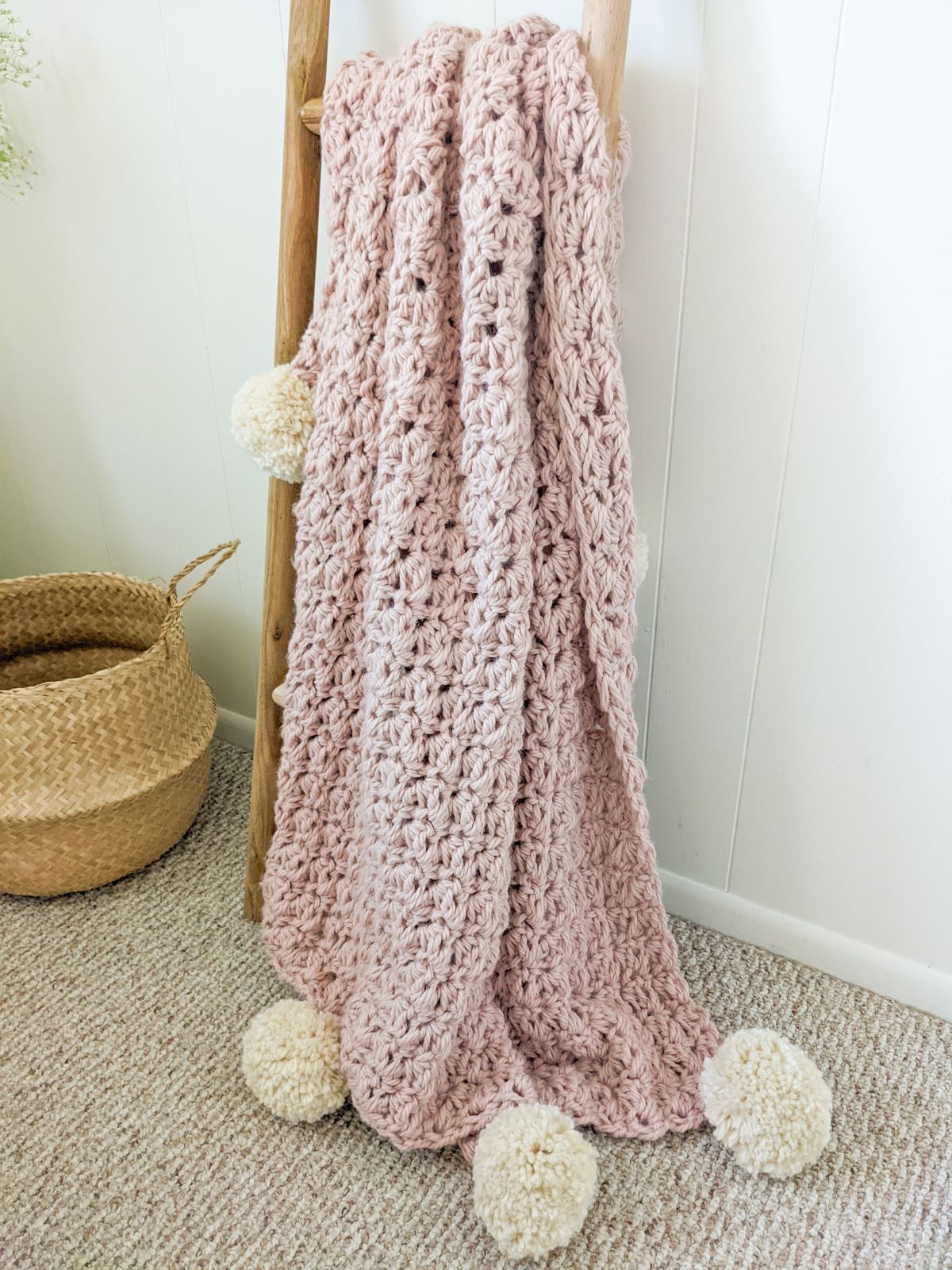 A light pink bulky weight crochet blanket on a decorative ladder with a belly basket.