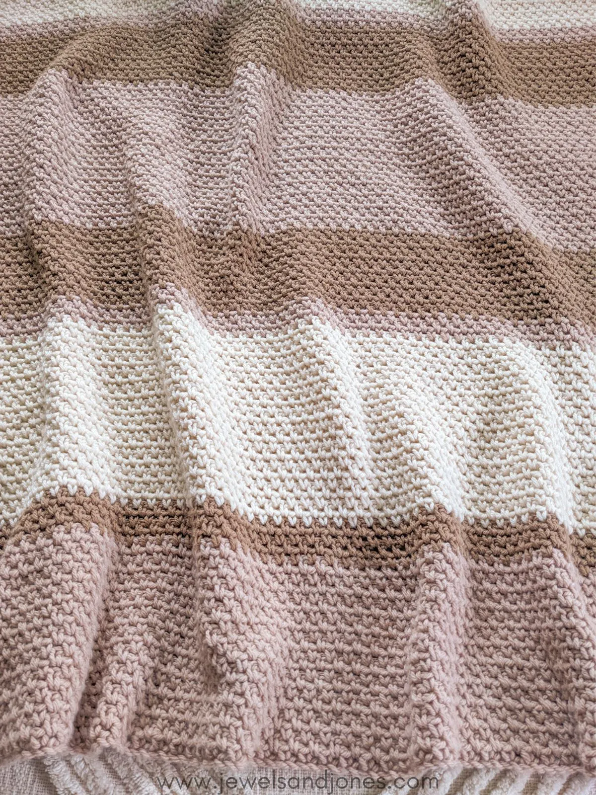 A crochet blanket that has 3 different colors, white, brown, and light pink. 